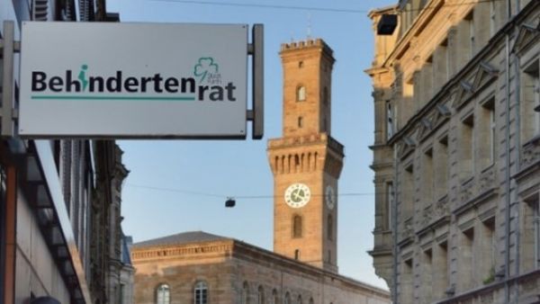 Der Fürther Rathausturm im Hintergrund wird von der späten Nachmittagssonne angestrahlt. Im Vordergrund ragt von links das Schild des Behindertenrats ins Bild. Dieses ist an der modernen Fassade des technischen Rathauses über dem Eingang der Hirschenstraße 2a angebracht. Bildnachweis: Verena Satzinger