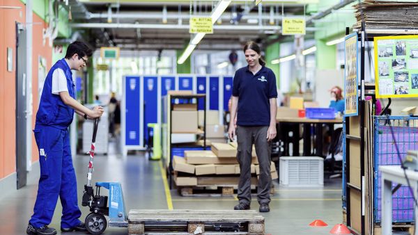 Arbeit im Lager der Dambacher Werkstätten in Fürth. Eine Person in Arbeitskleidung nimmt mit einem Hubwagen eine Europalette auf. Eine weitere Person leitet die Arbeit an. Bildnachweis: Lebenshilfe Fürth e.V.