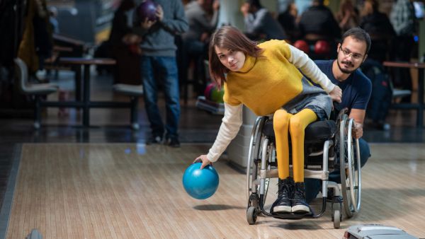 Bowling im Rollstuhl ist mit einer Persönlichen Assistenzkraft kein Problem. Eine junge Frau in sonnengelber Kleidung neigt sich zum Wurf der Bowlingkugel rechts aus ihrem Rollstuhl. Eine männliche Assistenz in der Hocke stabilisiert den Rollstuhl dabei von hinten. Er neigt seinen Kopf in die Gegenrichtung, um das Ergebnis des Wurfes zu beobachten. Bildnachweis: Adobe Stock