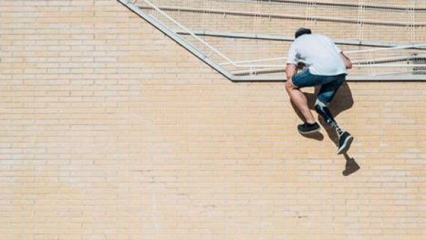 Ein Parcoursläufer mit Unterschenkelprothese kürzt den Weg über eine Treppe ab, indem er die senkrechte Seitenwand davon mit Griff ans Geländer erklimmt. Er ist schon fast über die Brüstung. Im Schatten, den er wirft, erkennt man erst richtig die Umrisse des rechten Beins und damit die Prothese. Bildnachweis: Westend61